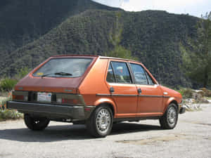 橙色復古 Fiat Strada 停在碎石路上，靠近綠色山丘和樹木。 — 飛雅特 壁紙