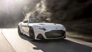 白色Aston Martin DBS在日落時分的森林道路上疾馳。 — 奧斯頓馬丁 壁紙
