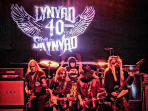 Lynyrd Skynyrd 樂團在舞台上現場表演，身後有發光的翅膀標誌。 — 音樂 壁紙