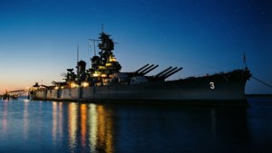 一艘戰艦夜間停泊，燈光在星空下映照在水面上。