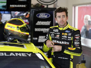 NASCAR車手Ryan Blaney身穿賽車裝備，站在車庫內他的黃色賽車旁。 — 賽車 壁紙