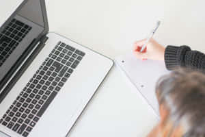 Person writing in notebook next to laptop on white desk — 讀書 壁紙