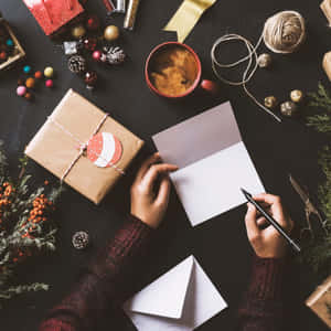Hands writing a holiday card next to a wrapped gift and festive decorations. — 1:1 壁紙