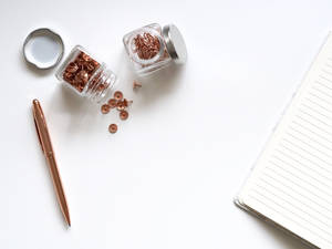 Rose gold stationery items on a white desk surface including push pins and pen. — 玫瑰金 壁紙