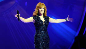 歌手 Reba McEntire 在舞台上穿著黑色亮片連衣裙，雙臂伸展表演。 — 音樂 壁紙