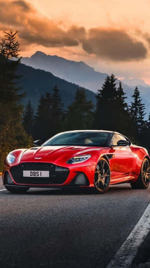 日落時分山路上的紅色Aston Martin DBS1，伴隨著松樹和山峰。 — 奧斯頓馬丁 壁紙