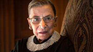 Ruth Bader Ginsburg 穿著帶蕾絲領的司法長袍，坐在華麗的椅子上。 — 露絲·拜德·金斯伯格 壁紙