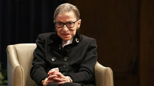 Ruth Bader Ginsburg 坐在米色扶手椅上，穿著黑色外套和眼鏡。 — 露絲·拜德·金斯伯格 壁紙