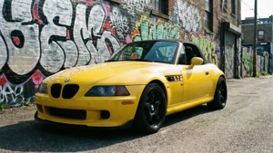 黃色BMW Z3敞篷車停泊在日落時分、旁邊有塗鴉磚牆的街道上。