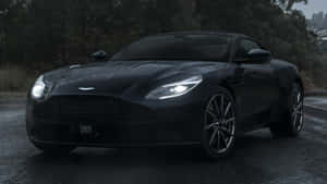 夜間濕路上黑色Aston Martin DB11，背景有城市和樹木。 — 奧斯頓馬丁 壁紙