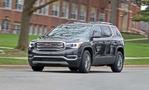 深灰色 GMC Acadia SUV 在磚砌建築和樹木附近的城市街道上行駛。 — Steam Deck 壁紙