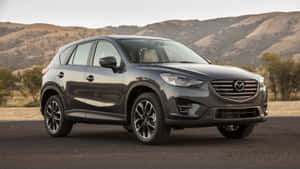 深灰色Mazda CX-5 SUV停在鋪砌道路上，靠近乾燥山丘和樹木。 — 馬自達 壁紙