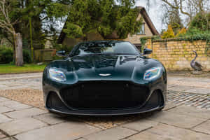 深綠色 Aston Martin 跑車停在石頭車道上，靠近石牆和樹木。 — 奧斯頓馬丁 壁紙