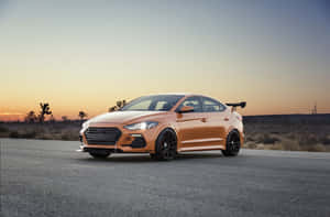 橙色Hyundai Elantra N在日落時分的沙漠公路上，背景有約書亞樹。 — Surface Pro 壁紙
