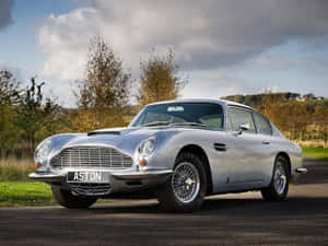 銀色經典Aston Martin DB5停在有樹木和多雲天空的道路上。 — 奧斯頓馬丁 壁紙