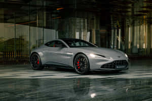 銀色Aston Martin Vantage停放在玻璃建築附近的潮濕路面上 — MacBook Pro M2 壁紙