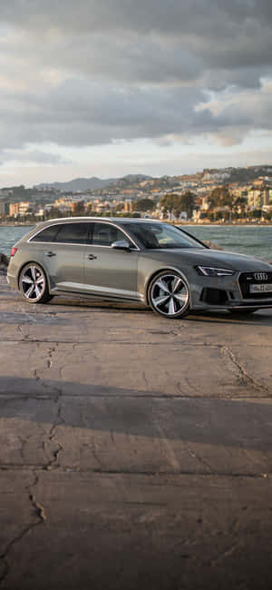 銀色 Audi 旅行車停在海邊混凝土上，背景有城市和水。 — iPhone 14 壁紙