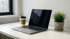 銀色筆記型電腦、咖啡杯和盆栽植物在窗邊的白色桌子上。