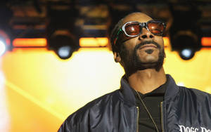 Snoop Dogg 戴著太陽眼鏡和飛行員夾克在舞台上，黃色燈光下。 — 史努比狗狗 壁紙