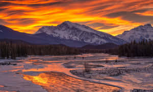 在鮮豔橙色夕陽天空下閃耀的雪山和河流。 — 日落 壁紙