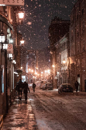 夜晚城市街道上飄雪，有發光的路燈和行人。 — 暮光 壁紙