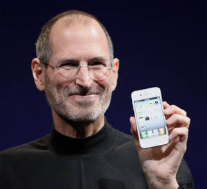 Steve Jobs 在舞台上帶著溫暖的笑容手持 iPhone。 — 蘋果 壁紙