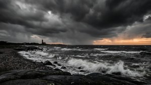 暴風雨的海浪拍打岩石海岸，烏雲密布，遠方燈塔在日落時發光。