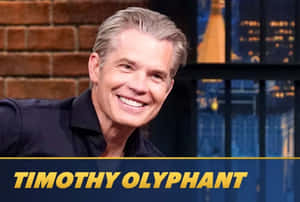 演員 Timothy Olyphant 在工作室中微笑，背景是磚牆和城市燈光。 — 達斯·尼希勒斯 壁紙