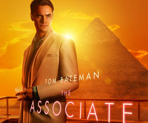 Tom Bateman 身穿米色西裝，在夕陽下的金字塔前拿著飲料。