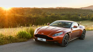 銅色Aston Martin停在夕陽下的道路上，背後有山丘和樹木。 — 奧斯頓馬丁 壁紙