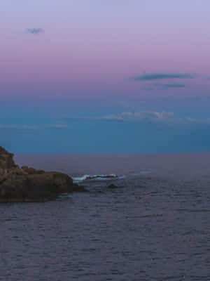 紫色和藍色的天空籠罩著平靜的海洋，海浪在黃昏時拍打岩石海岸。 — Kindle 壁紙