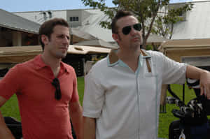 兩名身穿高爾夫球服裝的男子站在綠色球場上的高爾夫球車旁。 — MacBook Air M3 壁紙