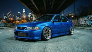 藍色改裝轎車夜間停放在城市高架橋下，帶有塗鴉牆和天際線。