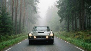 復古綠色跑車在迷霧森林道路上行駛，兩旁有高大松樹。