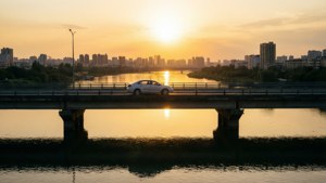 白色汽車在金色日落時分跨越河流上的橋樑，背景是城市天際線。