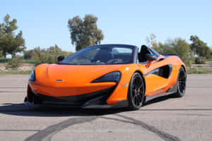 橙色McLaren 570S Spider敞篷車停放在柏油路上，背景有樹木和藍天。 — MacBook Pro 14 壁紙