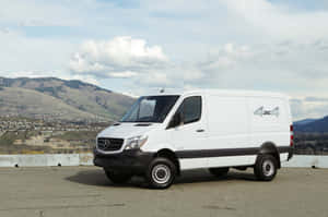 白色 Mercedes-Benz Sprinter 4x4 廂型車停放在風景優美的觀景點，背景有丘陵和城鎮。 — Surface Laptop 壁紙