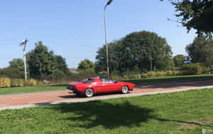 紅色經典跑車在陽光明媚的日子裡靠近綠樹和草地行駛。 — Steam Deck OLED 壁紙