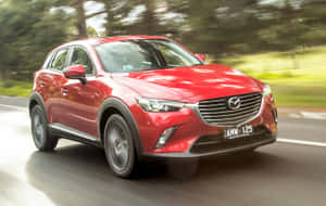 紅色Mazda CX-3 SUV在陽光明媚的道路上快速行駛，周圍有綠色樹木。 — 馬自達 壁紙