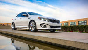 白色Kia Optima轎車停泊在水景旁邊，背景有建築物。 — 起亞 壁紙
