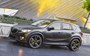 消光灰色馬自達CX-5 SUV，金色輪圈，在黃昏時停在一座現代建築前。 — 馬自達 壁紙