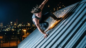 男子在夜間攀爬金屬屋頂，火花在城市燈光上方飛舞。