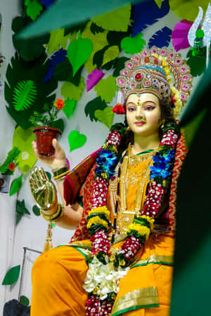 一尊裝飾過的印度教女神雕像，手持盆栽植物，背景為紙葉。 — Maa Durga 壁紙