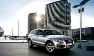 銀色 Audi Q7 SUV 停放在海濱附近，有帆船和現代建築。 — 奧迪 GIF 壁紙
