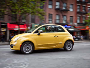 一輛黃色的Fiat 500在街道上行駛，背景建築物模糊。 — 飛雅特 壁紙