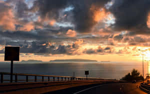 日落時分的沿海高速公路，帶有戲劇性的橙色雲彩和遠處的島嶼 — Steam Deck OLED 壁紙