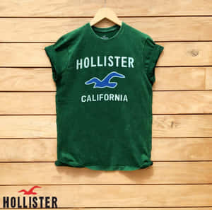 綠色Hollister California T恤掛在木衣架上，背景為木牆。 — 1:1 壁紙