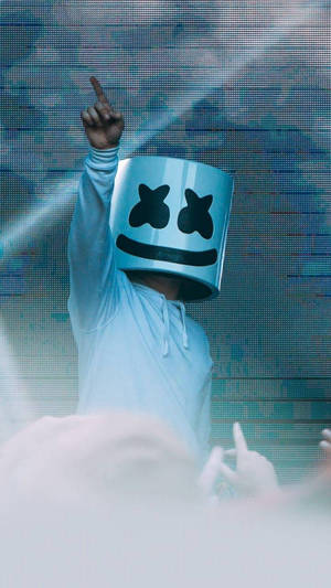 DJ戴著標誌性白色頭盔在舞台上向上指，背後有發光的燈光。 — Marshmello iPhone 壁紙