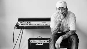 戴帽子的刺青音樂家坐在單色工作室中的鍵盤和 Marshall 擴音器旁邊。 — 美學 Mac 壁紙