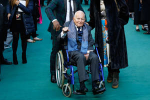 一位坐在輪椅上的長者男士在藍綠色地毯上微笑，周圍有人群。 — 好萊塢 壁紙